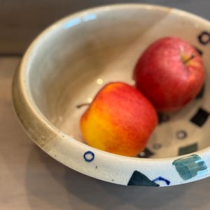 May include: A ceramic bowl with a beige interior and a decorative rim. The rim features blue, black, and green geometric designs. Two red apples with yellow highlights rest inside the bowl, creating a still life composition.