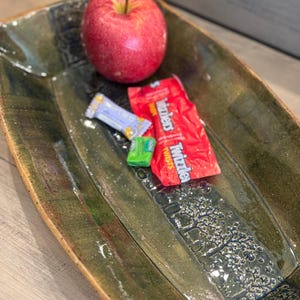 May include: An olive green ceramic serving dish with a rectangular shape and a textured tree design. It contains a red apple and wrapped sweets, displaying a rustic aesthetic.
