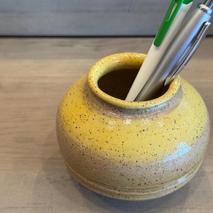 Puede incluir: Un pequeño portalápices redondo de cerámica amarilla con un diseño moteado. Dos bolígrafos, uno verde y blanco, el otro plateado, están dentro. El portalápices tiene una abertura ancha y una superficie ligeramente texturizada.