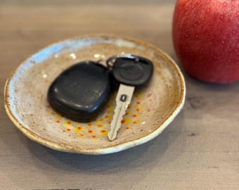 May include: A small, round, ceramic dish with a speckled, tan glaze holds a black car key fob and a silver key. A red apple sits to the right. The dish has a yellow and red dotted pattern.