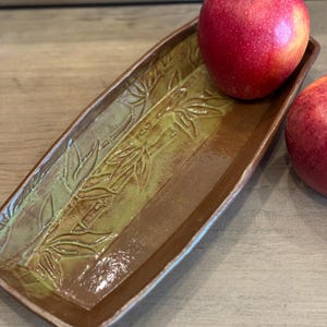 Peut inclure: Un plat de service rectangulaire en céramique avec un motif de feuilles de bambou dans des tons de vert et de marron. Deux pommes rouges reposent sur le plat. Le plat mesure environ 30 cm de long et 13 cm de large.