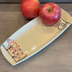 May include: A rectangular ceramic serving platter with a cream-colored interior and white trim. The platter is decorated with colorful tree and circle designs. Two red apples rest on the wooden surface next to the platter.