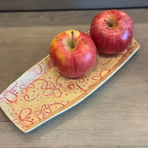 May include: Two red apples sit on a rectangular, yellow ceramic tray. The tray is decorated with red floral patterns. The apples are a vibrant red with yellow accents.