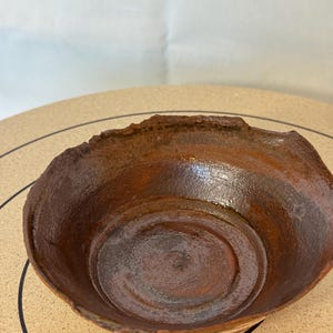 May include: A handmade, rustic ceramic bowl with a dark brown, textured exterior and a lighter brown interior. The bowl has an irregular, slightly chipped rim and a circular base. The bowl is likely used for serving food or as a decorative piece.