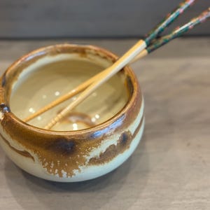 May include: A ceramic bowl with a cream interior and a brown, textured exterior. Two wooden chopsticks with green and floral designs rest across the bowl, suggesting a dining or serving theme.