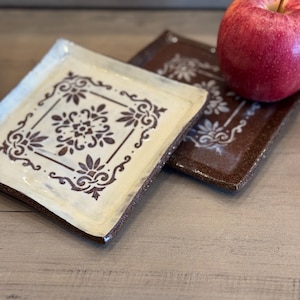 Könnte beinhalten: Zwei quadratische Keramikplatten mit floralen Designs. Eine Platte ist cremefarben mit braunen Akzenten, die andere ist braun mit cremefarbenen Akzenten. Ein roter Apfel liegt auf der braunen Platte. Die Platten haben ein rustikales, handgemachtes Aussehen.