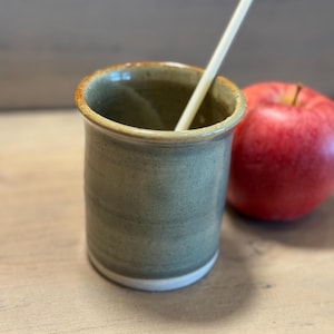 May include: A handmade, olive green ceramic tumbler with a white base and a yellow straw inside. A red apple rests beside the tumbler on a wooden surface. The tumbler has a rustic, textured appearance.
