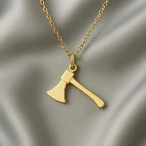 Pode incluir: Um colar com pingente de machado dourado. O pingente de machado é uma miniatura detalhada, suspensa por uma corrente delicada. O colar é exibido em um fundo cinza suave, destacando o design do pingente.