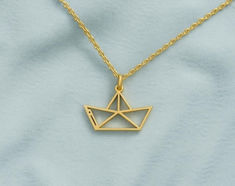May include: A gold-colored necklace featuring a pendant shaped like a paper boat. The pendant is a geometric design with clean lines, suspended from a delicate chain. The necklace is displayed against a light blue background.