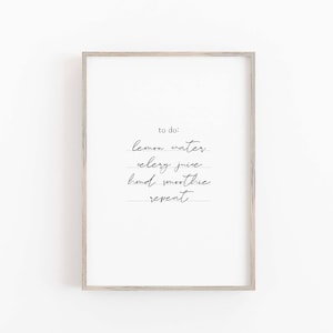 May include: A framed print with a white background and handwritten text. The text reads "to do: lemon water, celery juice, hand smoothie, repeat" in a delicate, cursive font. The frame is a light, natural wood color.