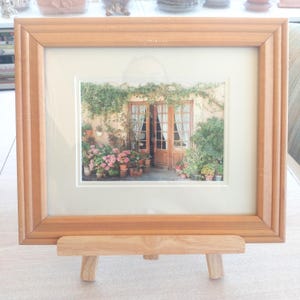 May include: A framed photograph of a courtyard with wooden doors, surrounded by potted plants and flowers. The frame is light brown and rests on a small wooden easel.