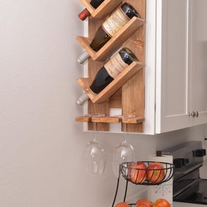 May include: Wooden wine rack holding three bottles of wine, mounted on a white wall. Two wine glasses hang below the rack. A two-tiered black wire basket holds apples and oranges.
