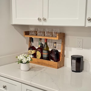 May include: A wooden bar shelf with six glasses and four bottles of liquor. The shelf is mounted under a white cabinet. A small white flower pot and a black candle are on the counter.