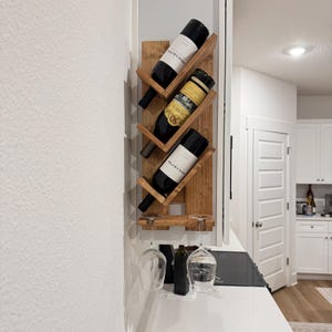 May include: A wooden wine rack with three angled shelves holding wine bottles. Below, a small shelf holds two wine glasses and a bottle. The rack is mounted on a white wall, showcasing a rustic home decor element.