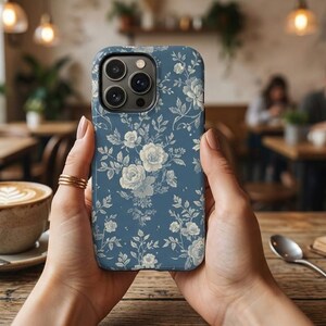 Puede incluir: Una funda para smartphone con fondo azul polvoriento y un patrón floral repetitivo de rosas y hojas blancas. La funda tiene un recorte para un sistema de cámara de tres lentes en la parte posterior, mostrando un diseño botánico detallado.