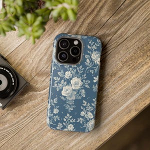 May include: A blue phone case with a floral pattern featuring white roses and leaves. The case is on a wooden surface, with a camera and plant visible in the background. The floral design covers the entire case.