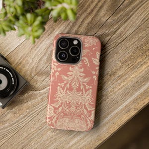 May include: A coral-colored phone case with a cream-colored floral damask pattern. The case is on a wooden surface, with a camera and plant in the background. The case has a glossy finish and a protective design.