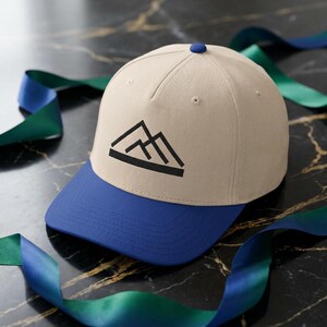 May include: A beige baseball cap with a blue brim and a black mountain logo. The cap has a blue button on top. The cap is made of a soft material.