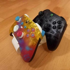 Può includere: Due controller Xbox. Uno è nero con un motivo mimetico, l'altro è un design personalizzato con una parte superiore gialla e un design a spruzzi di vernice colorati in rosso, blu e viola. I controller sono su una superficie di legno.