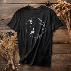 May include: Black t-shirt featuring a circular design with a silhouette of a woman's profile. The design is in shades of white and gray, with flowing hair and a detailed outline. The shirt is made of a soft material.
