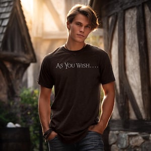 May include: A dark brown t-shirt with the text "As You Wish...." in white. The short-sleeved shirt is worn by a person standing outdoors. The person is wearing blue jeans and has their hands in their pockets.