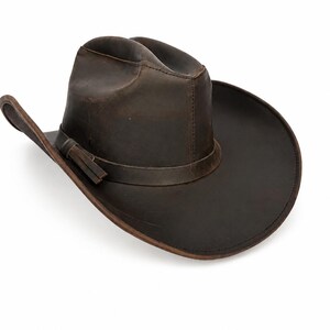 May include: A dark brown leather cowboy hat with a distressed finish, featuring a pinched crown, a wide curved brim, and a matching leather hat band with a small side detail. The hat has visible stitching along the crown and brim edges.