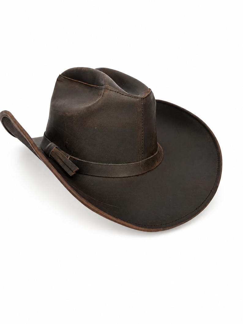 May include: A dark brown leather cowboy hat with a distressed finish, featuring a pinched crown, a wide curved brim, and a matching leather hat band with a small side detail. The hat has visible stitching along the crown and brim edges.