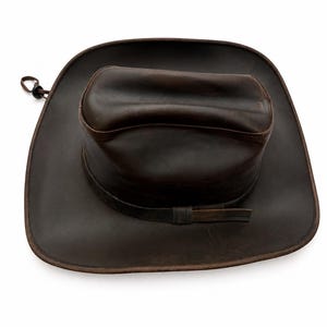 May include: A dark brown leather cowboy hat with a wide brim and a creased crown. The hat features a matching leather band around the base of the crown and a thin cord with a small black plastic toggle attached to the side of the brim.