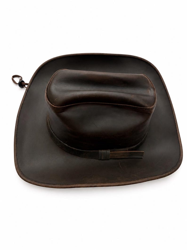 May include: A dark brown leather cowboy hat with a wide brim and a creased crown. The hat features a matching leather band around the base of the crown and a thin cord with a small black plastic toggle attached to the side of the brim.