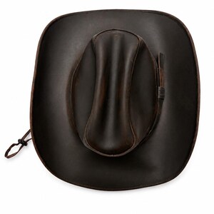 May include: A dark brown leather cowboy hat with a wide brim and a creased crown. The hat features a matching leather band around the base of the crown and a thin adjustable chin strap with a small black plastic toggle attached to the side.