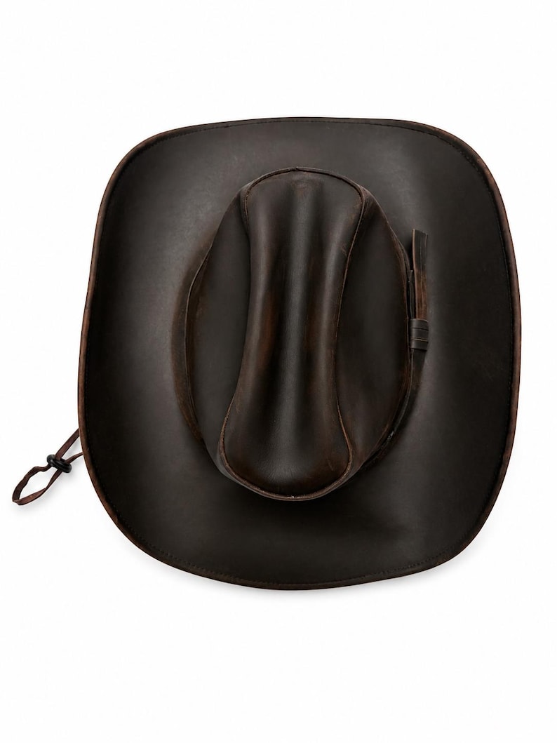 May include: A dark brown leather cowboy hat with a wide brim and a creased crown. The hat features a matching leather band around the base of the crown and a thin adjustable chin strap with a small black plastic toggle attached to the side.