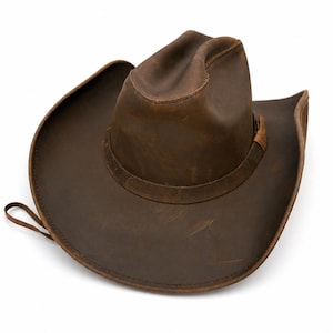 May include: A brown leather cowboy hat with a distressed finish, featuring a pinched crown, a wide curved brim, and a matching leather hat band. The material shows natural variations in color and texture, with visible stitching along the brim.