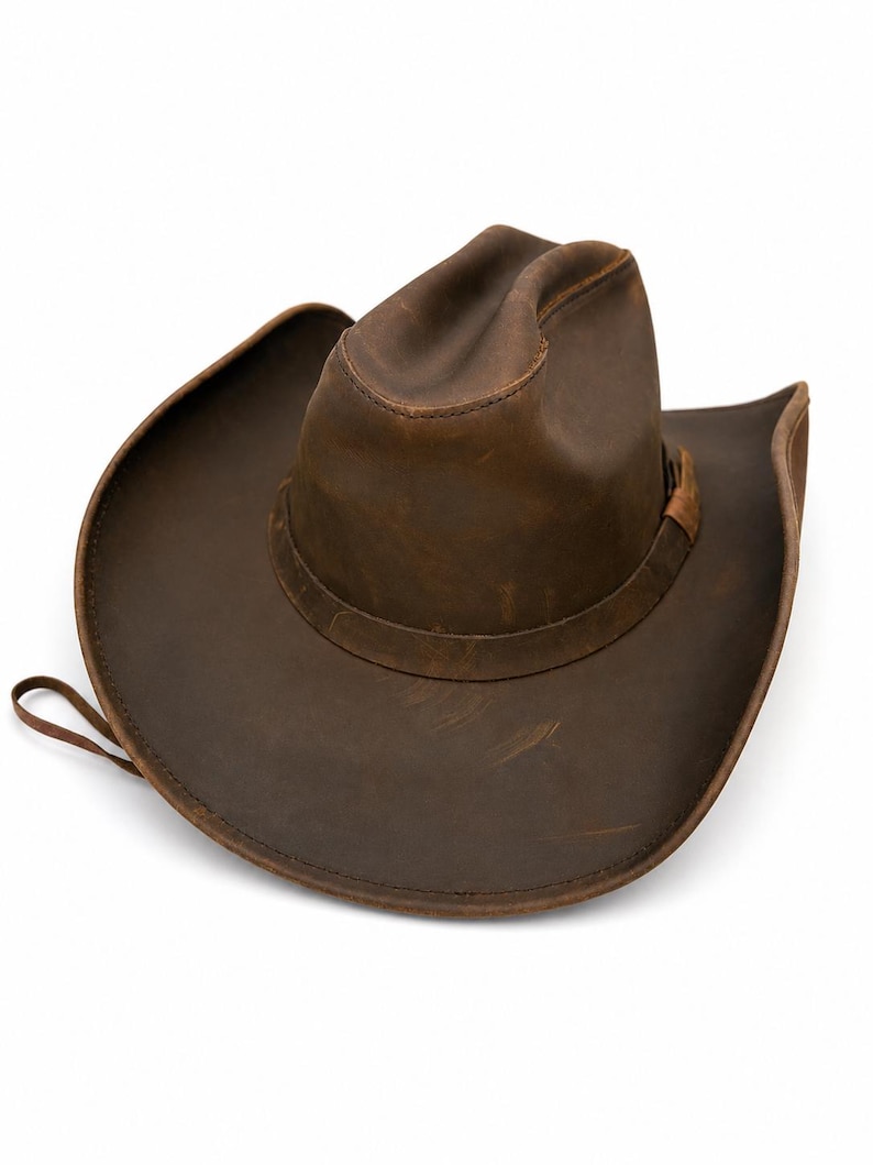 May include: A brown leather cowboy hat with a distressed finish, featuring a pinched crown, a wide curved brim, and a matching leather hat band. The material shows natural variations in color and texture, with visible stitching along the brim.