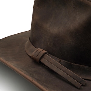 May include: A dark brown leather hat featuring a wide brim and a matching leather hat band. The band is wrapped around the base of the crown and secured with a simple knot, with two flat leather tails extending from the side of the hat.