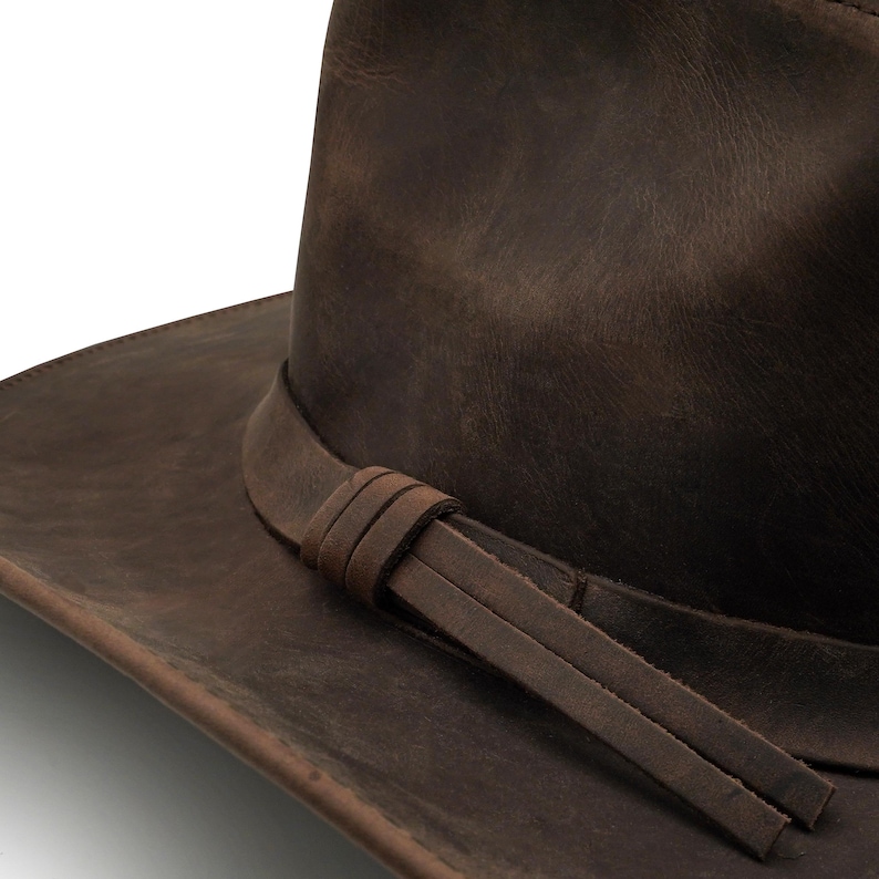May include: A dark brown leather hat featuring a wide brim and a matching leather hat band. The band is wrapped around the base of the crown and secured with a simple knot, with two flat leather tails extending from the side of the hat.