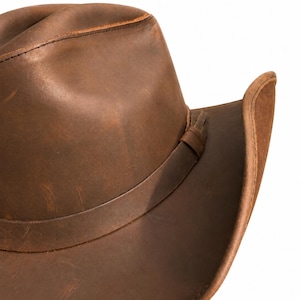 May include: A brown leather cowboy hat with a distressed finish, featuring a creased crown and a curved brim. A matching leather band wraps around the base of the crown, secured with a small leather loop detail on the side of the hat.