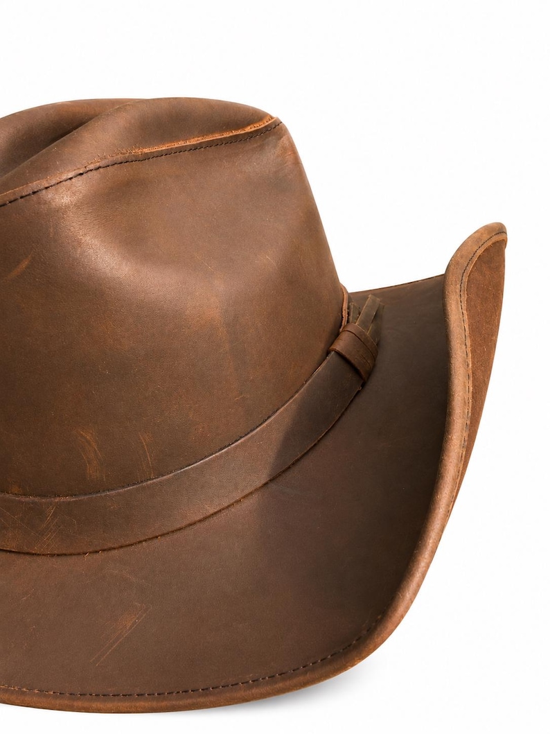 May include: A brown leather cowboy hat with a distressed finish, featuring a creased crown and a curved brim. A matching leather band wraps around the base of the crown, secured with a small leather loop detail on the side of the hat.