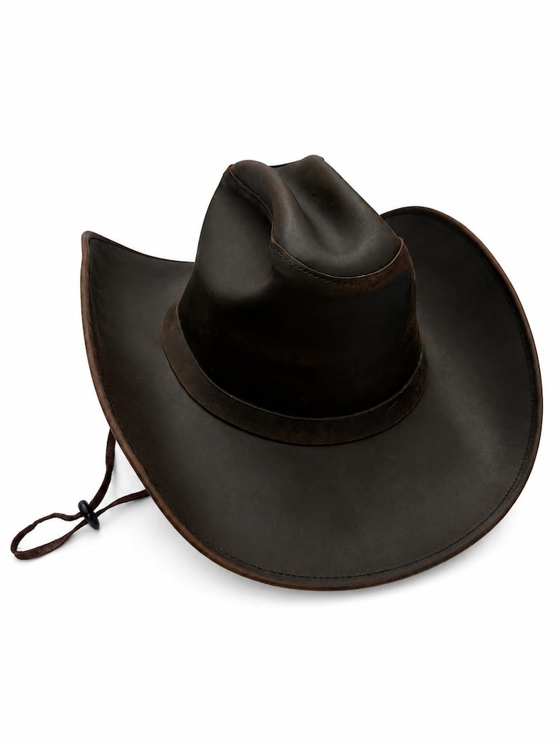 May include: A dark brown cowboy hat with a wide, curved brim and a pinched crown. The hat features a matching dark brown band around the base of the crown and a thin, adjustable brown chin cord with a small black plastic toggle for tightening.