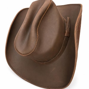 May include: A brown leather cowboy hat with a creased crown and a wide, curved brim. The hat features a smooth, matte finish with visible stitching along the edges of the brim and crown, and a thin leather band encircling the base of the crown.
