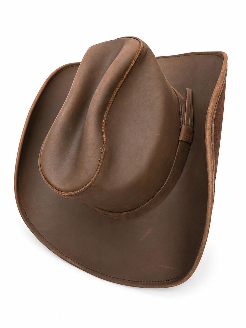 May include: A brown leather cowboy hat with a creased crown and a wide, curved brim. The hat features a smooth, matte finish with visible stitching along the edges of the brim and crown, and a thin leather band encircling the base of the crown.