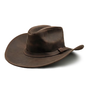 May include: A dark brown leather cowboy hat with a wide, curved brim and a creased crown. The hat features a matching leather band wrapped around the base of the crown, secured with a simple knot detail on the side, showcasing a smooth finish.