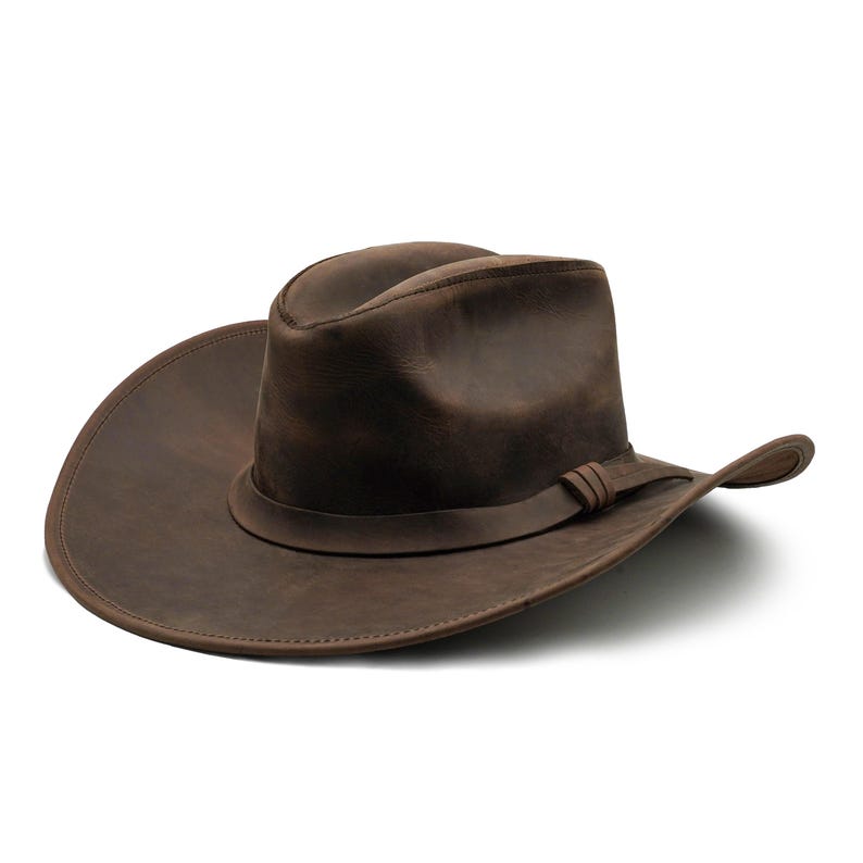 May include: A dark brown leather cowboy hat with a wide, curved brim and a creased crown. The hat features a matching leather band wrapped around the base of the crown, secured with a simple knot detail on the side, showcasing a smooth finish.