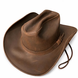 May include: A brown leather cowboy hat featuring a creased crown, a wide curved brim, and a matching leather hat band with a thin cord chin strap attached to the side. The surface of the hat displays natural variations in the leather texture.