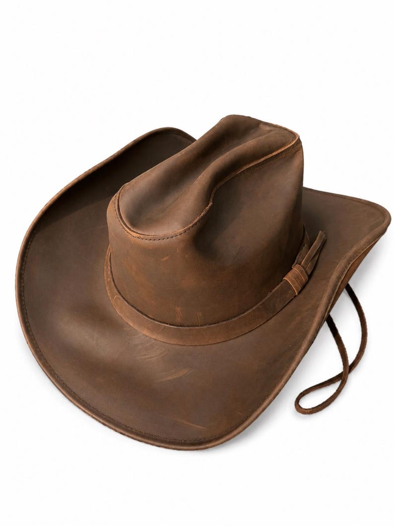 May include: A brown leather cowboy hat featuring a creased crown, a wide curved brim, and a matching leather hat band with a thin cord chin strap attached to the side. The surface of the hat displays natural variations in the leather texture.