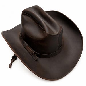 May include: A dark brown leather cowboy hat with a creased crown and a wide, curved brim. The hat features a matching leather hatband and a thin, adjustable leather chin cord with a small sliding toggle attached to the side of the brim.