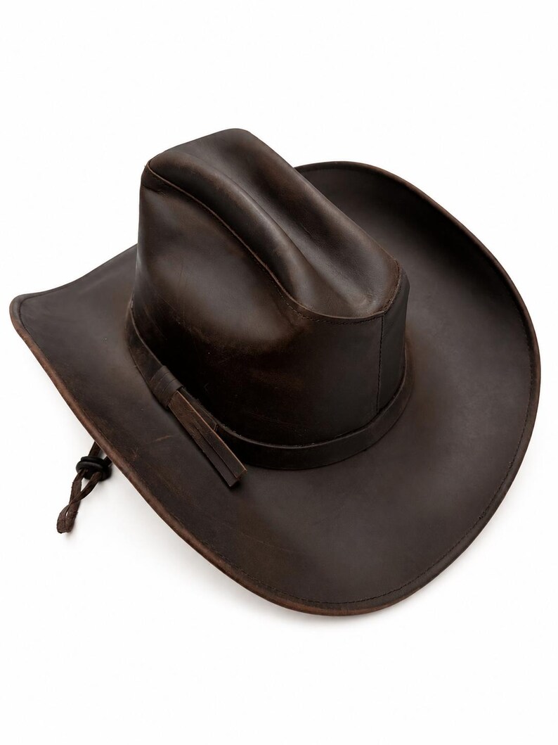 May include: A dark brown leather cowboy hat with a creased crown and a wide, curved brim. The hat features a matching leather hatband and a thin, adjustable leather chin cord with a small sliding toggle attached to the side of the brim.