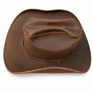 May include: A brown leather cowboy hat with a wide, curved brim and a creased crown. The hat features a smooth, matte finish with visible natural variations in the leather texture and a stitched band encircling the base of the crown.