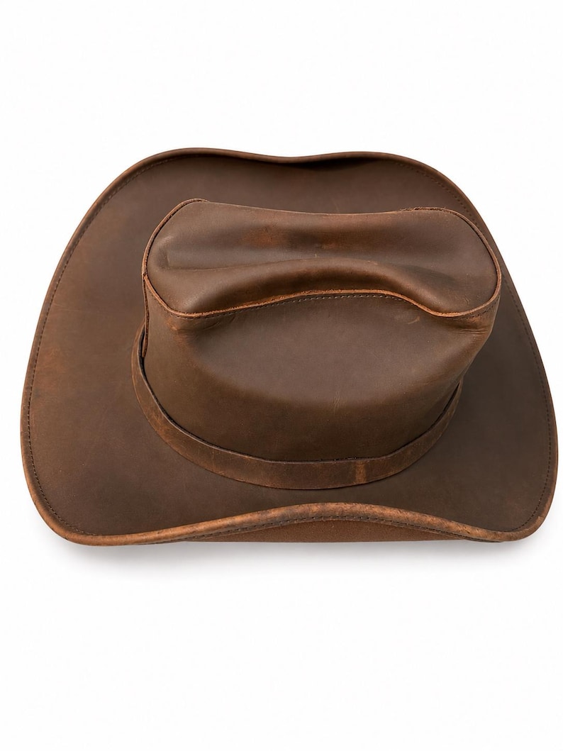 May include: A brown leather cowboy hat with a wide, curved brim and a creased crown. The hat features a smooth, matte finish with visible natural variations in the leather texture and a stitched band encircling the base of the crown.