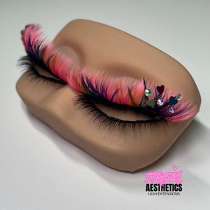 May include: A set of false eyelashes with pink, purple, and orange fibers applied to a mannequin eyelid. The lashes are decorated with small iridescent rhinestones and dark heart-shaped accents. The text ITGIRL AESTHETICS LASH EXTENSIONS is visible.