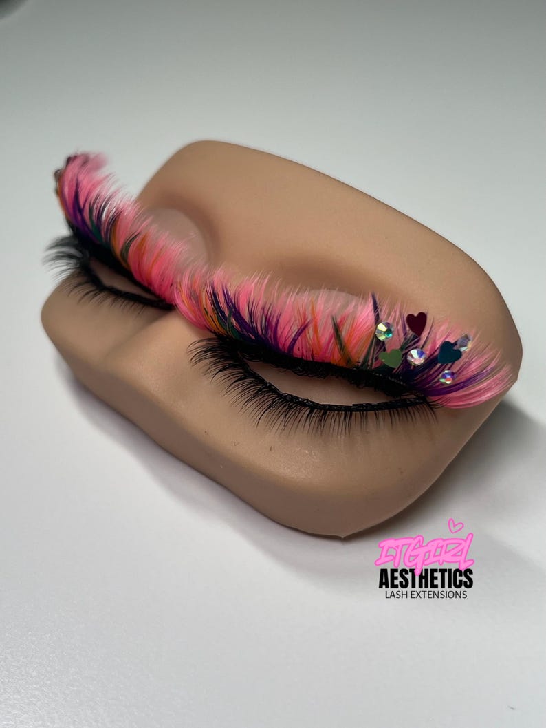 May include: A set of false eyelashes with pink, purple, and orange fibers applied to a mannequin eyelid. The lashes are decorated with small iridescent rhinestones and dark heart-shaped accents. The text ITGIRL AESTHETICS LASH EXTENSIONS is visible.